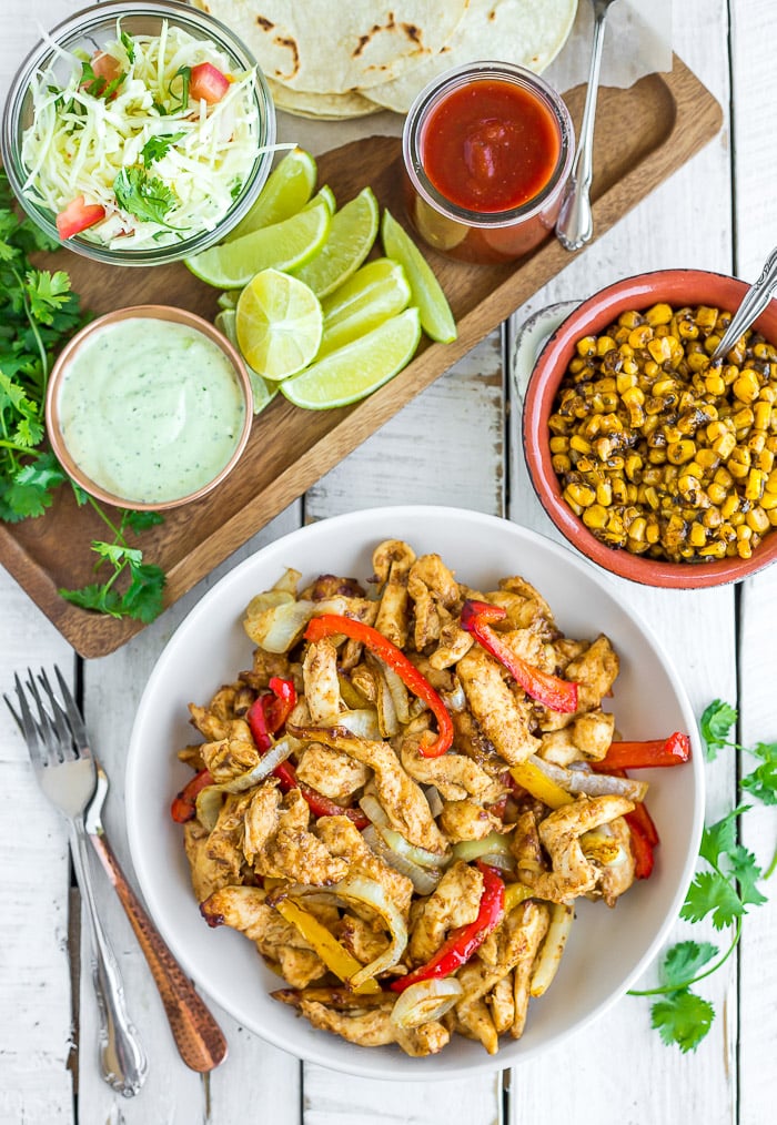 a bowl of sliced air fryer chicken fajitas with peppers and onions as well as sauce, limes, and other ingredients on a white wooden board.