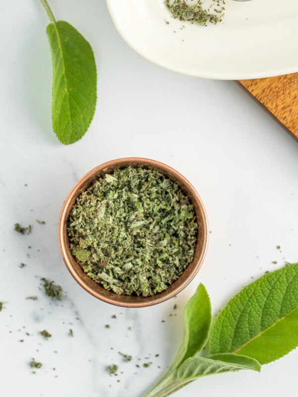 a small metal dish with dried sage in it, and fresh sage leaves on a white board.