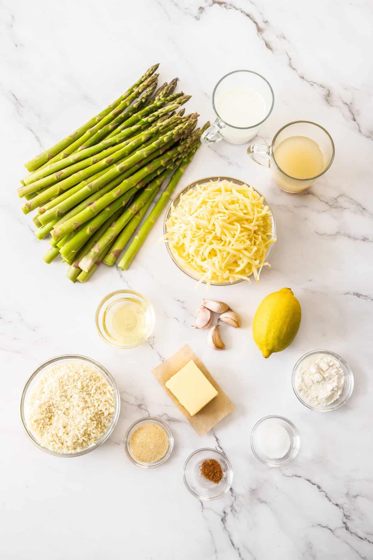 bowls of shredded cheese, panko, garlic, milk, butter, asparagus and other ingredients on a white board.