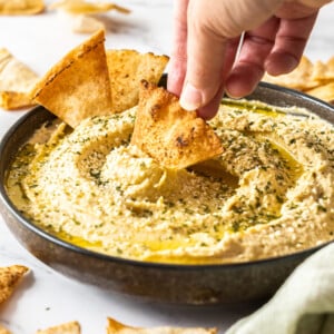 a hand with a pita chip dipping into a bowl of garlic hummus.