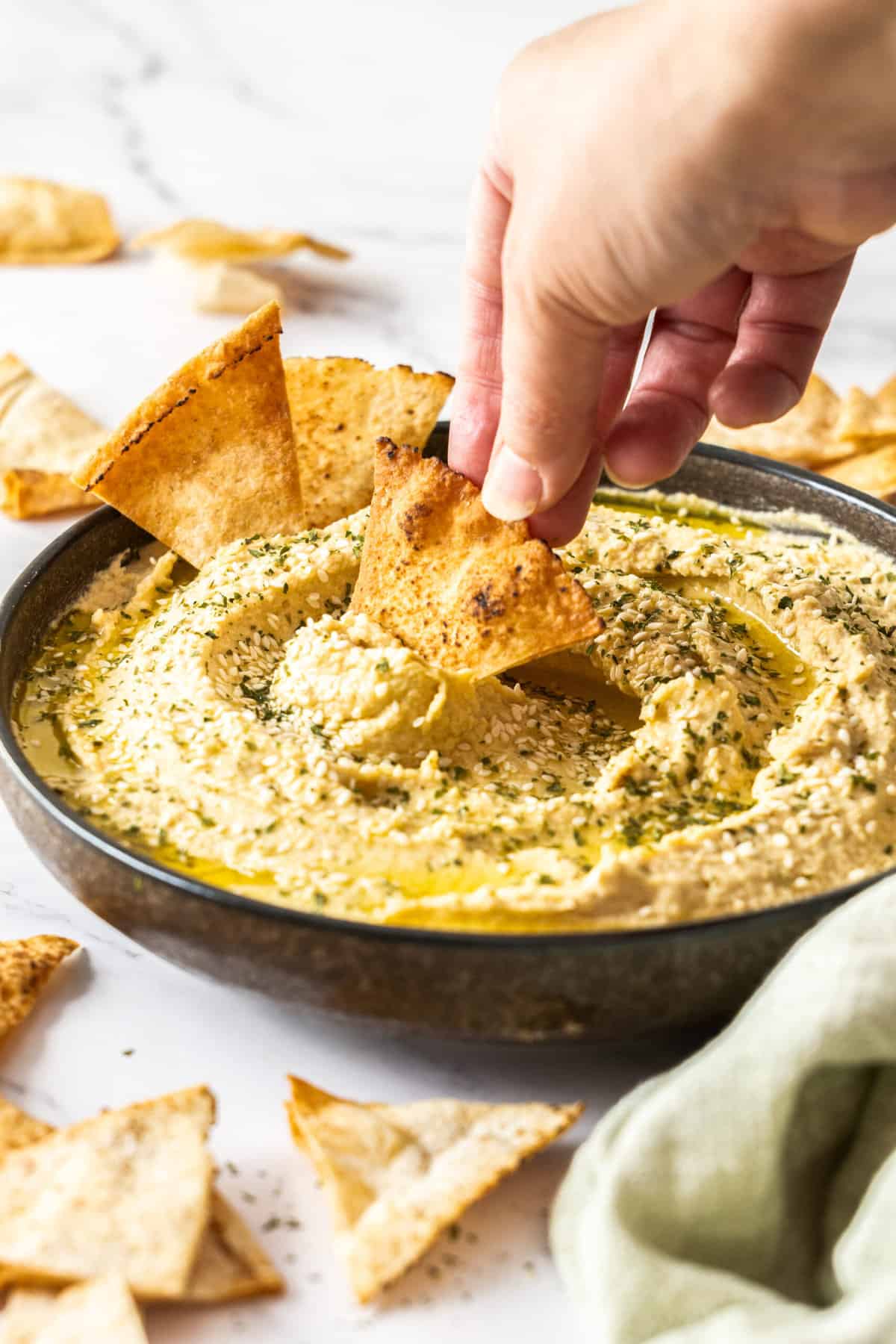 a hand with a pita chip dipping into a bowl of garlic hummus.