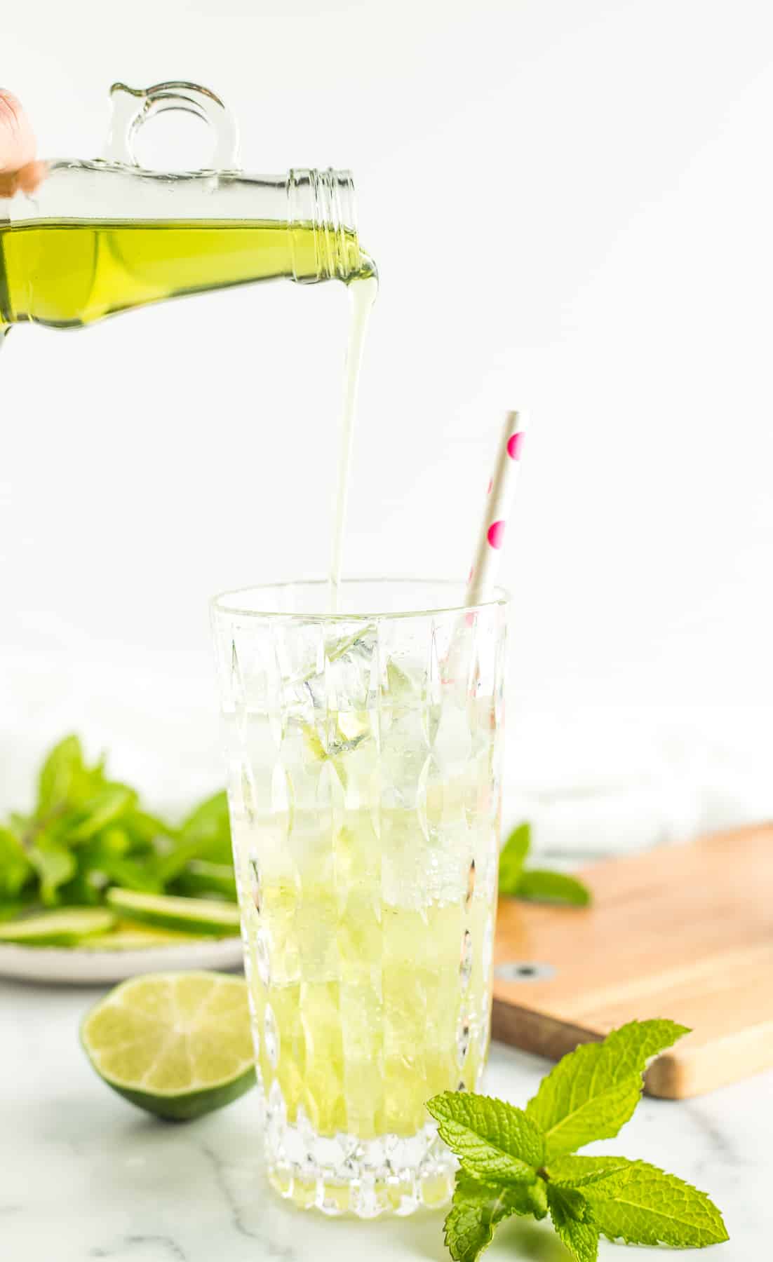 a bottle pouring green liquid into a glass filled with sparkling water, and ice cubes.