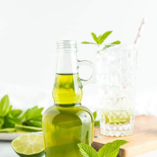 a glass bottle of green mint syrup with fresh mint and limes on a white board.