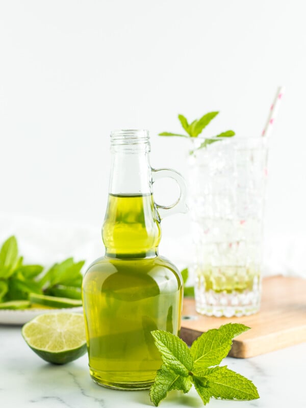 a glass bottle of green mint syrup with fresh mint and limes on a white board.