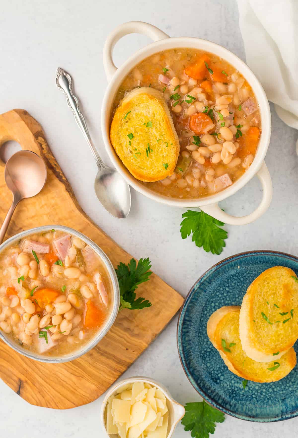 multiple bowls of beans and ham on a white surface with a dish of bread.