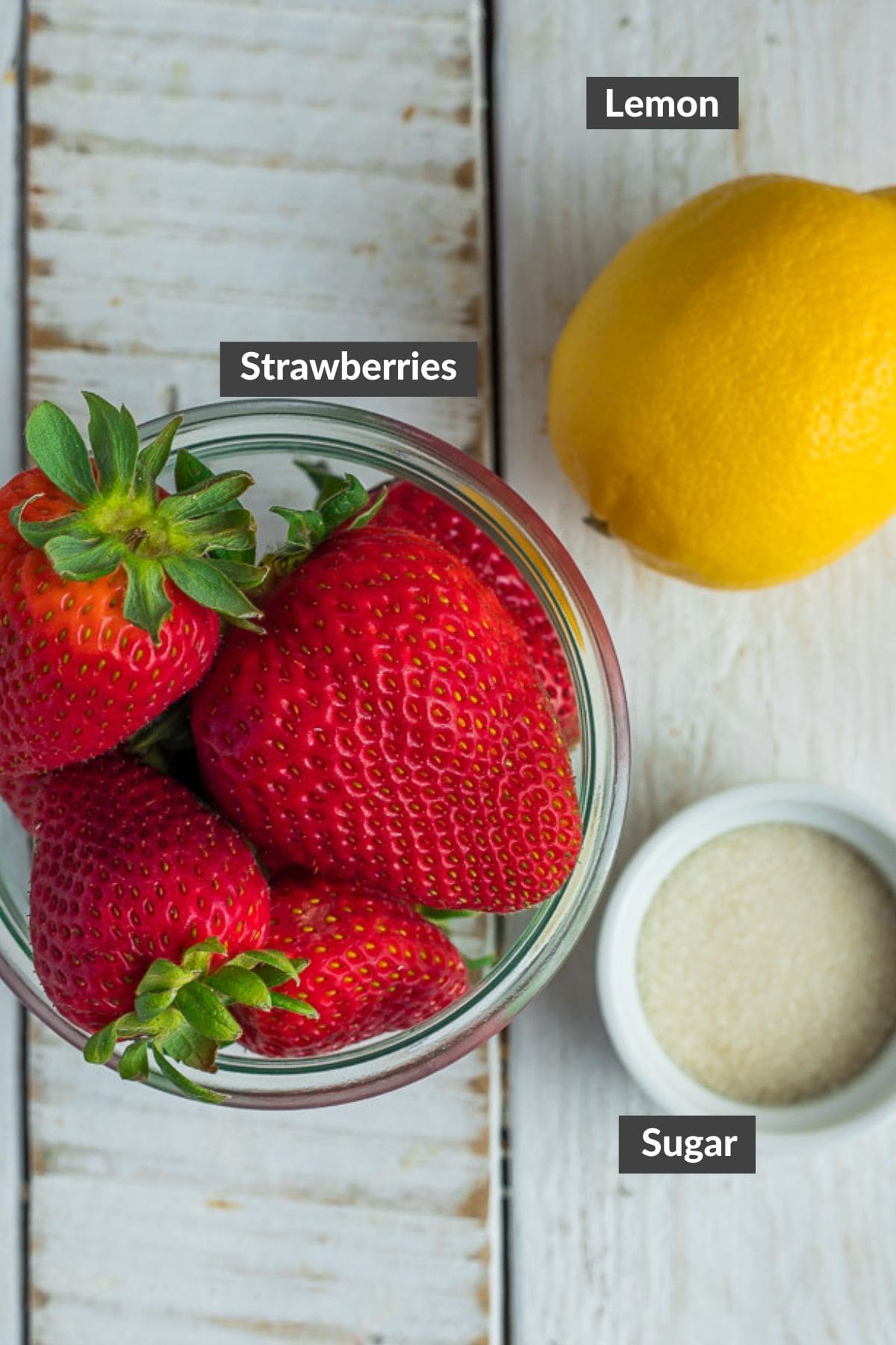 a glass bowl of strawberries, a white bowl of sugar, and a lemon on a wooden board.