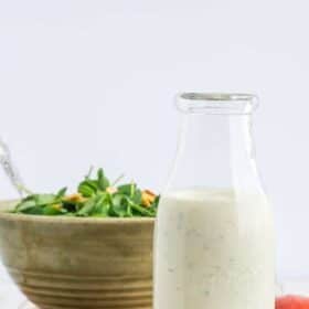 a glass bottle of yogurt ranch dressing on a cutting board with a bowl of salad.