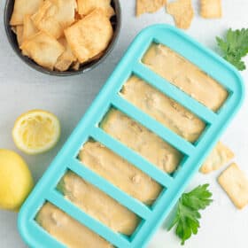 a teal freezer tray with hummus on a white board with a bowl of chips, lemons, and parsley.