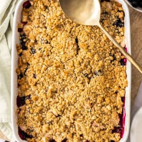 a white baking dish filled with apple and blueberry crumble with a gold spoon.