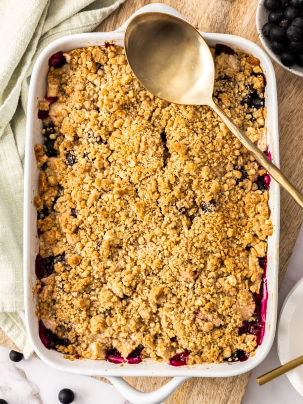 a white baking dish filled with apple and blueberry crumble with a gold spoon.