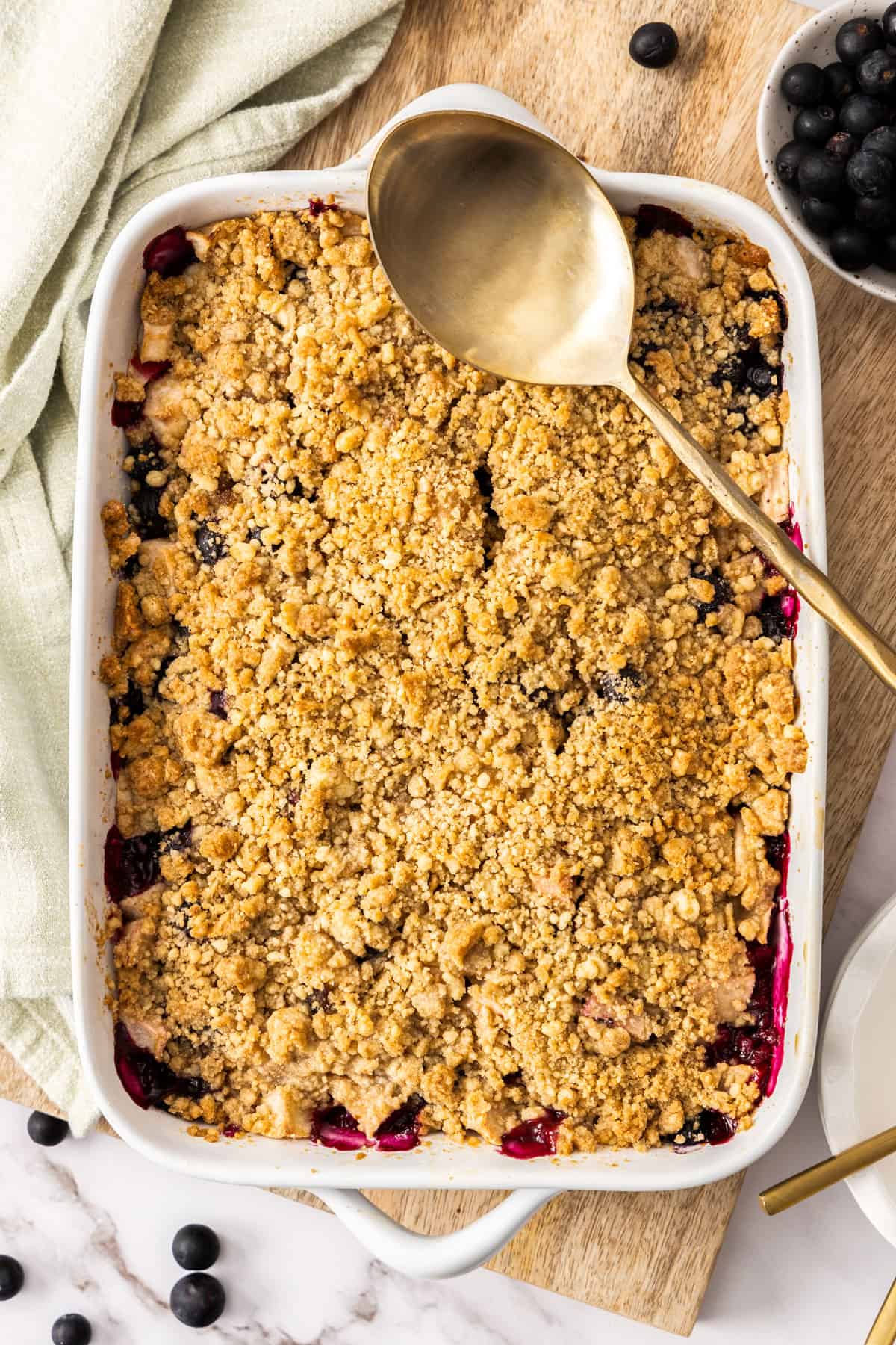 a white ceramic baking dish filled with an apple and blueberry crumble, topped with a gold serving spoon.