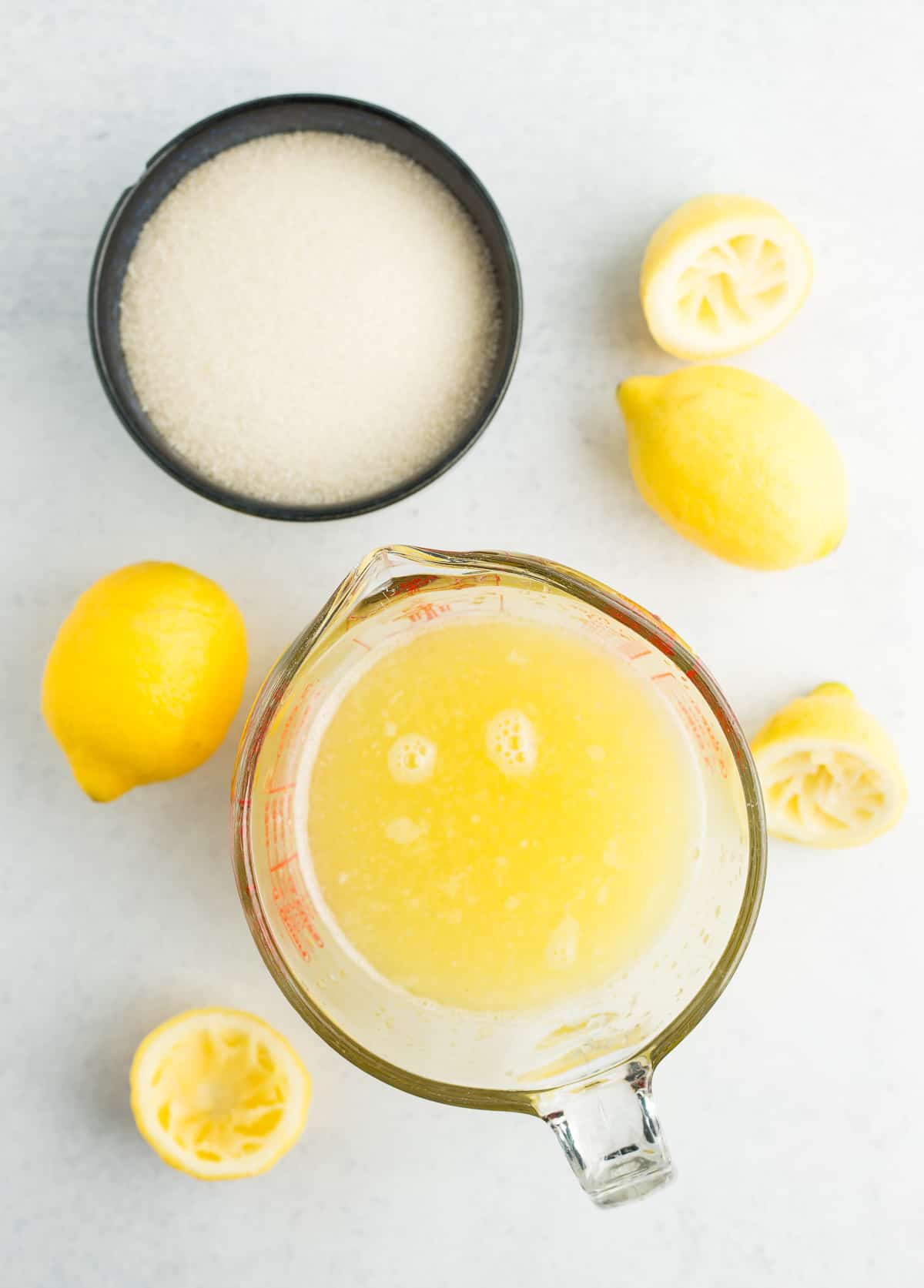 a measuring glass of lemon juice and a navy blue bowl of sugar with fresh lemons.
