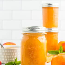 3 jars of peach preserves with fresh peaches and sprigs of mint on a white board.