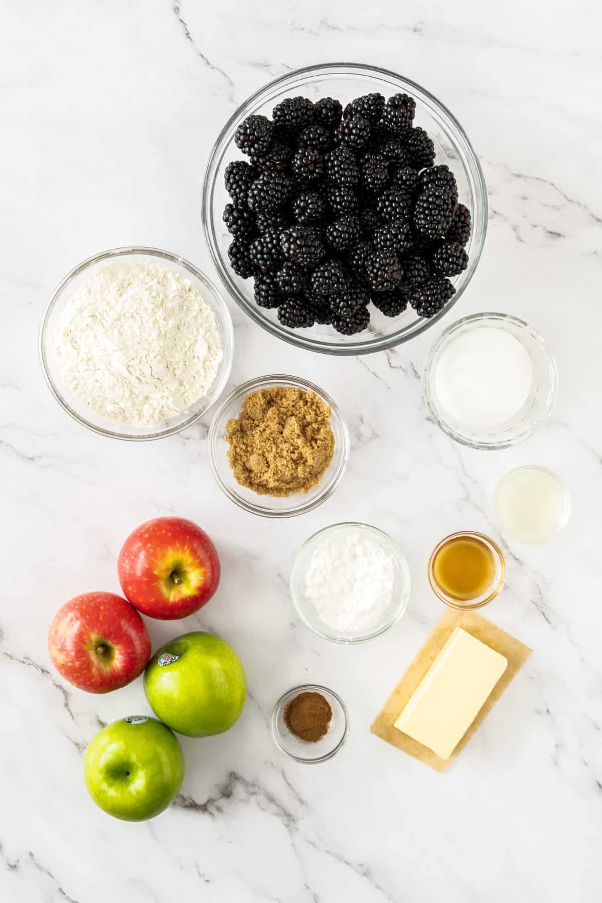 a bowl of blackberries, flour, brown sugar, butter, and apples on a marble board.