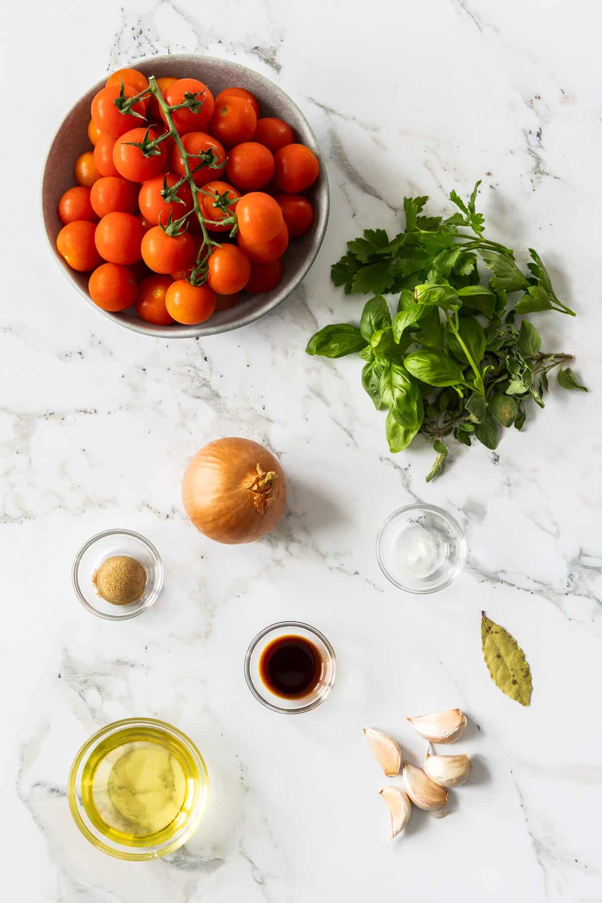 a bowl of cherry tomatoes, fresh herbs, garlic, onion, and other ingredients on a marble board.
