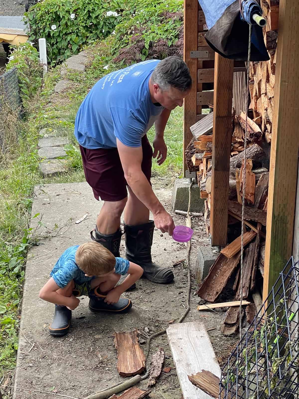 a boy and his dad both in blue shirts and boots in front of a woodshed.