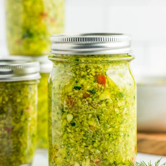 3 jars of zucchini relish on a marble board with a tile background.