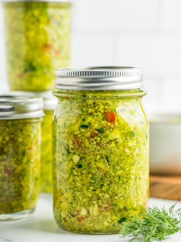 3 jars of zucchini relish on a marble board with a tile background.
