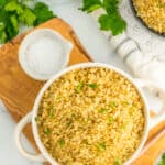 a white bowl with Instant Pot Quinoa topped with chopped parsley, sitting on a wooden board. There are small bowls of seasonings and parsley on a white marbled surface.