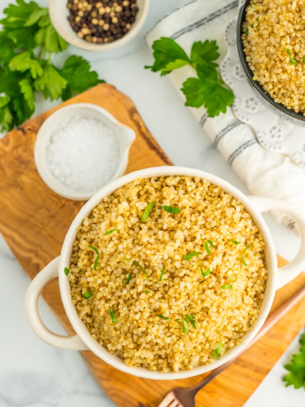 a white bowl with Instant Pot Quinoa topped with chopped parsley, sitting on a wooden board. There are small bowls of seasonings and parsley on a white marbled surface.