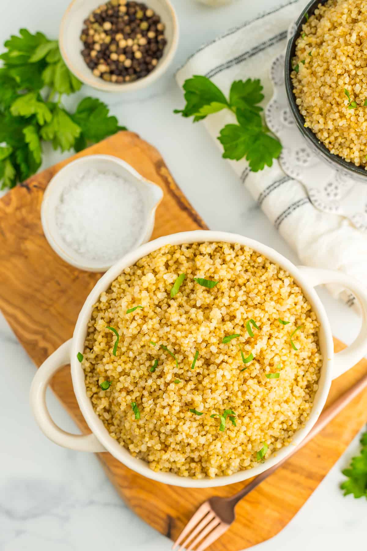 a white bowl with Instant Pot Quinoa topped with chopped parsley, sitting on a wooden board. There are small bowls of seasonings and parsley on a white marbled surface.