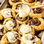 a spoonful of frosting hovering over a pan of cinnamon scrolls.