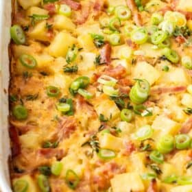a white baking dish with a ham and potato casserole topped with sliced green onions.