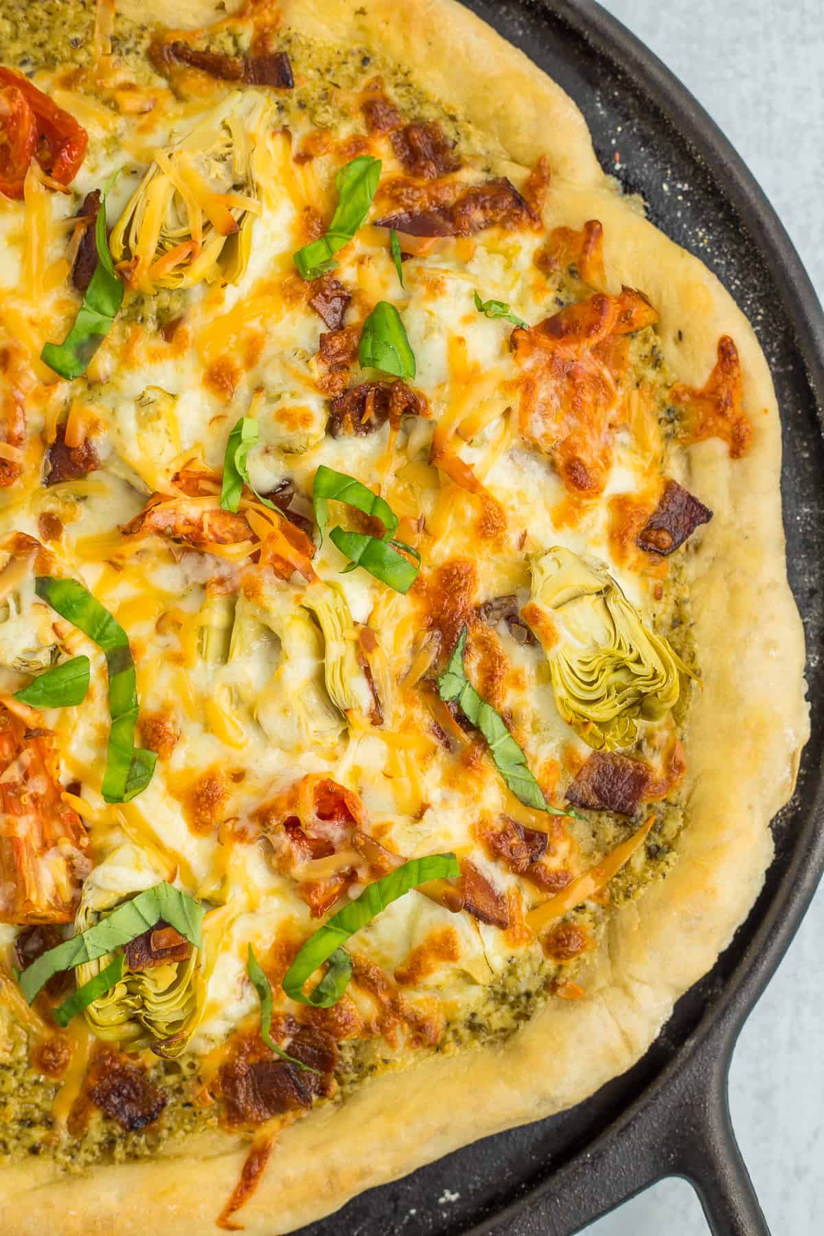 An homemade pizza on a cast iron pan topped with basil, artichoke hearts, and melted cheese.