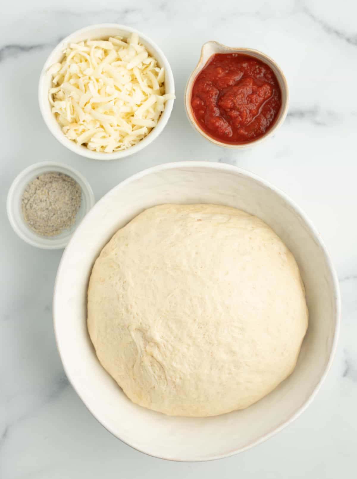 a bowl with dough, red sauce, shredded mozzarella, and cornmeal on a marbled board.