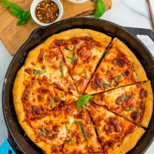 pizza in a skillet topped with basil, with two bowls above it full of shredded Parmesan and red pepper flakes.