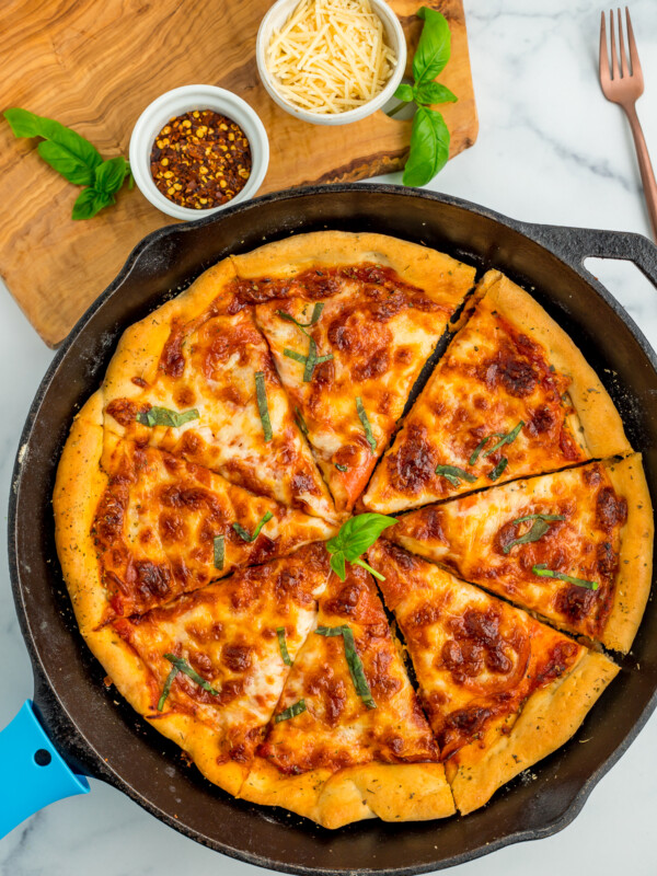 pizza in a skillet topped with basil, with two bowls above it full of shredded Parmesan and red pepper flakes.
