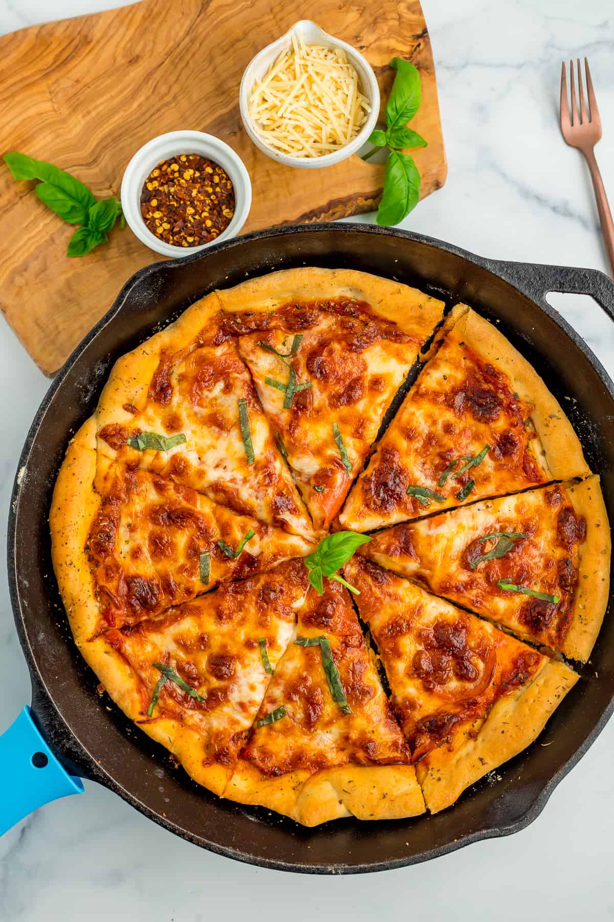 pizza in a skillet topped with basil, with two bowls above it full of shredded Parmesan and red pepper flakes.