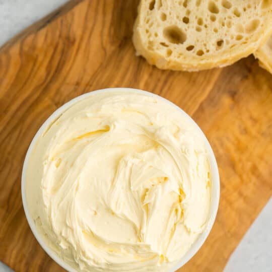 a white dish of whipped butter on a wooden board with slices of baguettes.