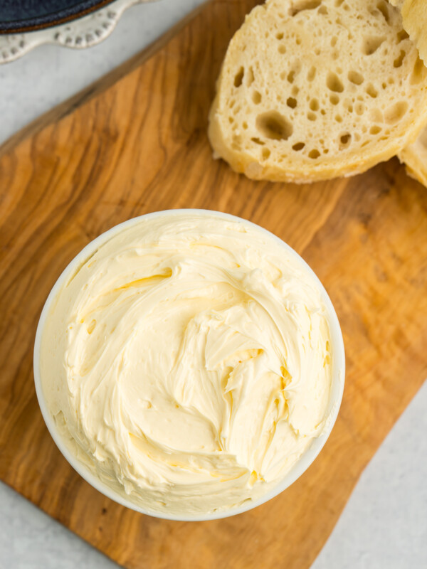 a white dish of whipped butter on a wooden board with slices of baguettes.