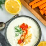 A dark blue bowl of whipped feta dip topped with fresh dill, chopped roasted tomatoes and garlic, with a lemon and veggies.