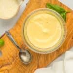 a glass dish with white pizza sauce on a wooden board with a small spoon, dish of Parmesan, and basil.