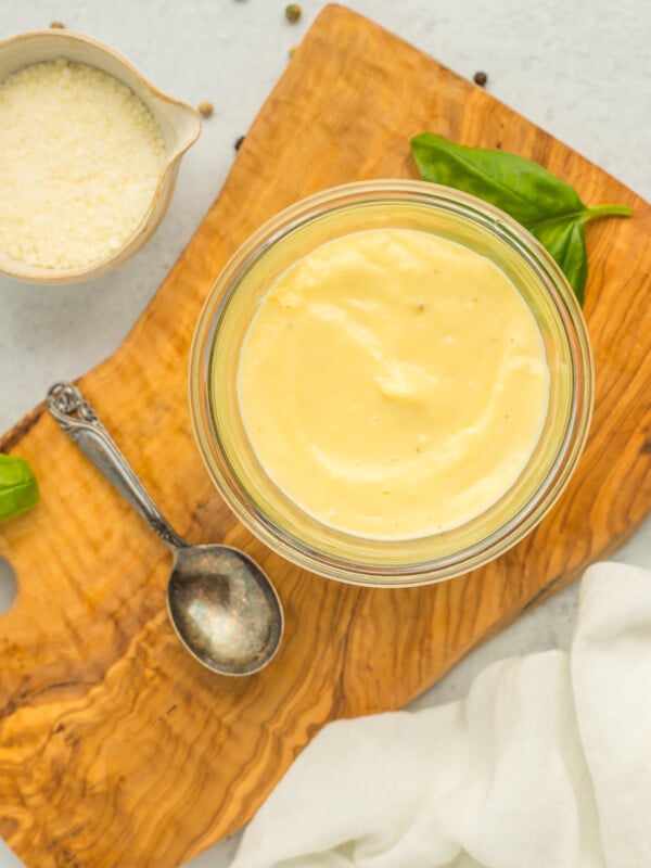 a glass dish with white pizza sauce on a wooden board with a small spoon, dish of Parmesan, and basil.