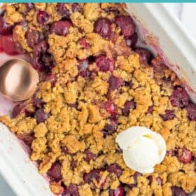 a white baking dish of cherry crumble with a scoop of ice cream on top, and a spoonful of crumble missing.