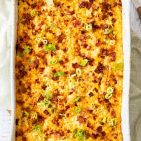 A hashbrown breakfast casserole topped with bacon and sliced green onions in a white baking dish.