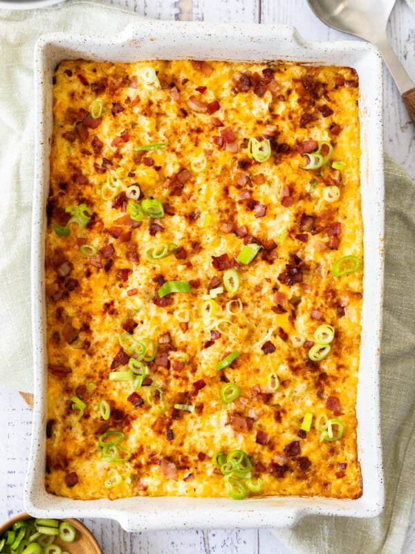 A hashbrown breakfast casserole topped with bacon and sliced green onions in a white baking dish.