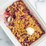 a white baking dish of cherry crumble with a scoop of ice cream on top, and a spoonful of crumble missing.