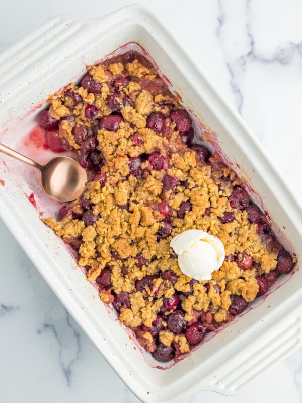 a white baking dish of cherry crumble with a scoop of ice cream on top, and a spoonful of crumble missing.