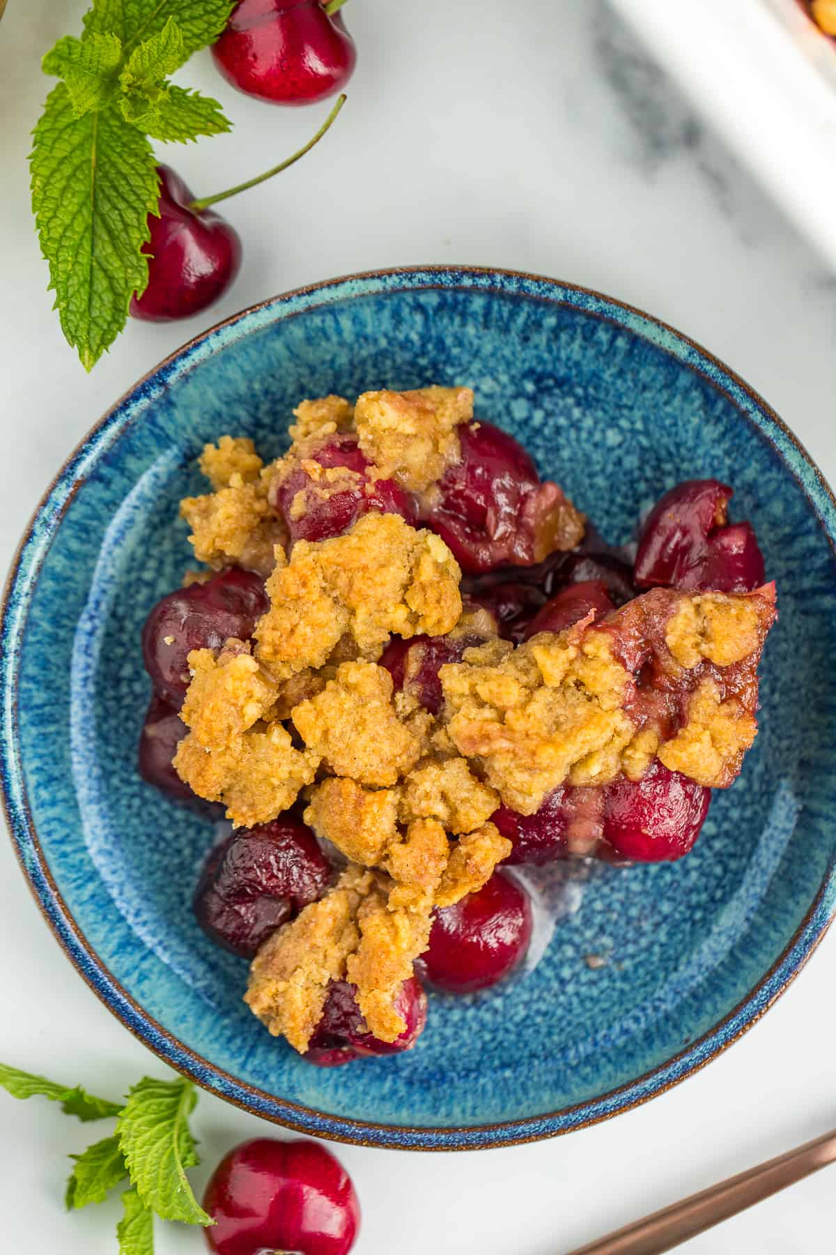 a blue dish with a serving of cherry crisp. There are fresh cherries and mint on a grey board with the plate.