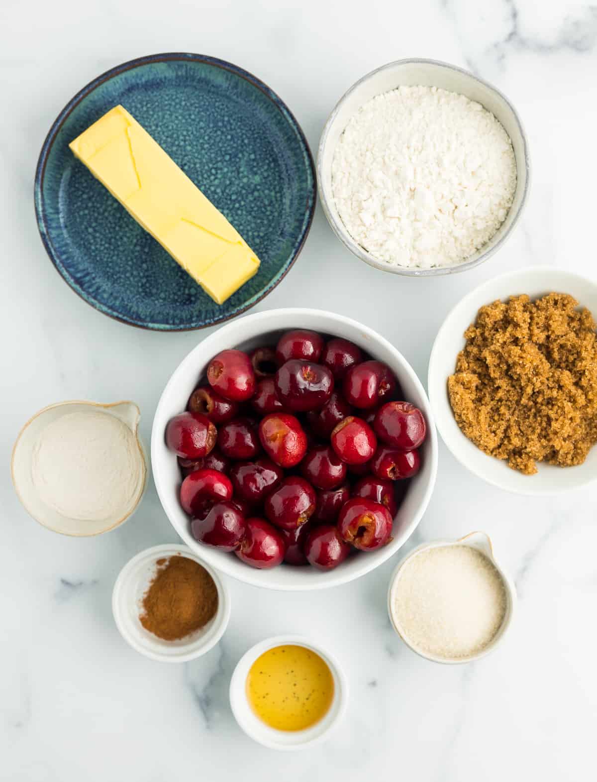 a plate of butter, a bowl of cherries, flour, sugar, and other ingredients on a marbled board.