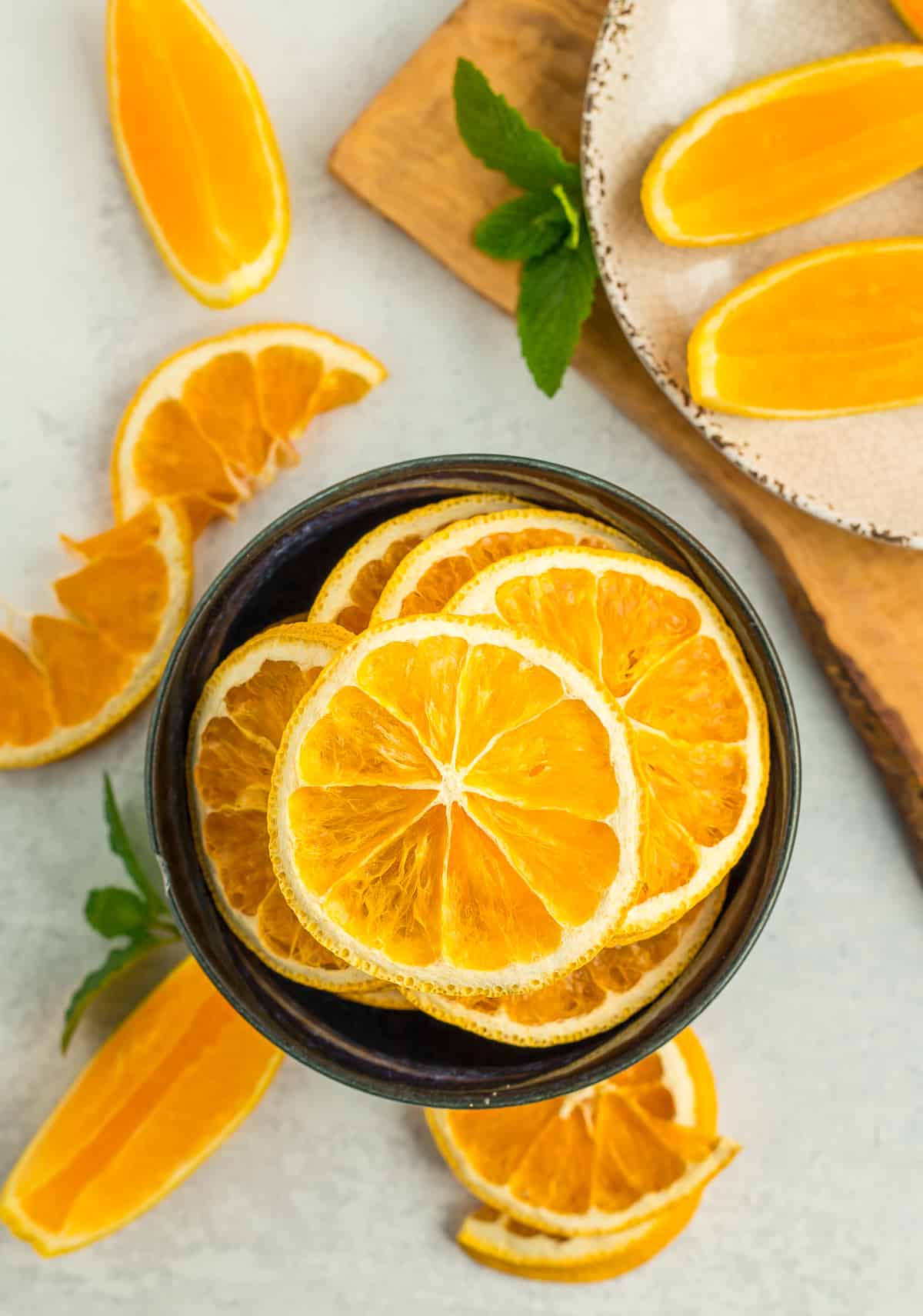 a blue bowl filled with dried orange slices, with dried and fresh orange slices on a grey board.