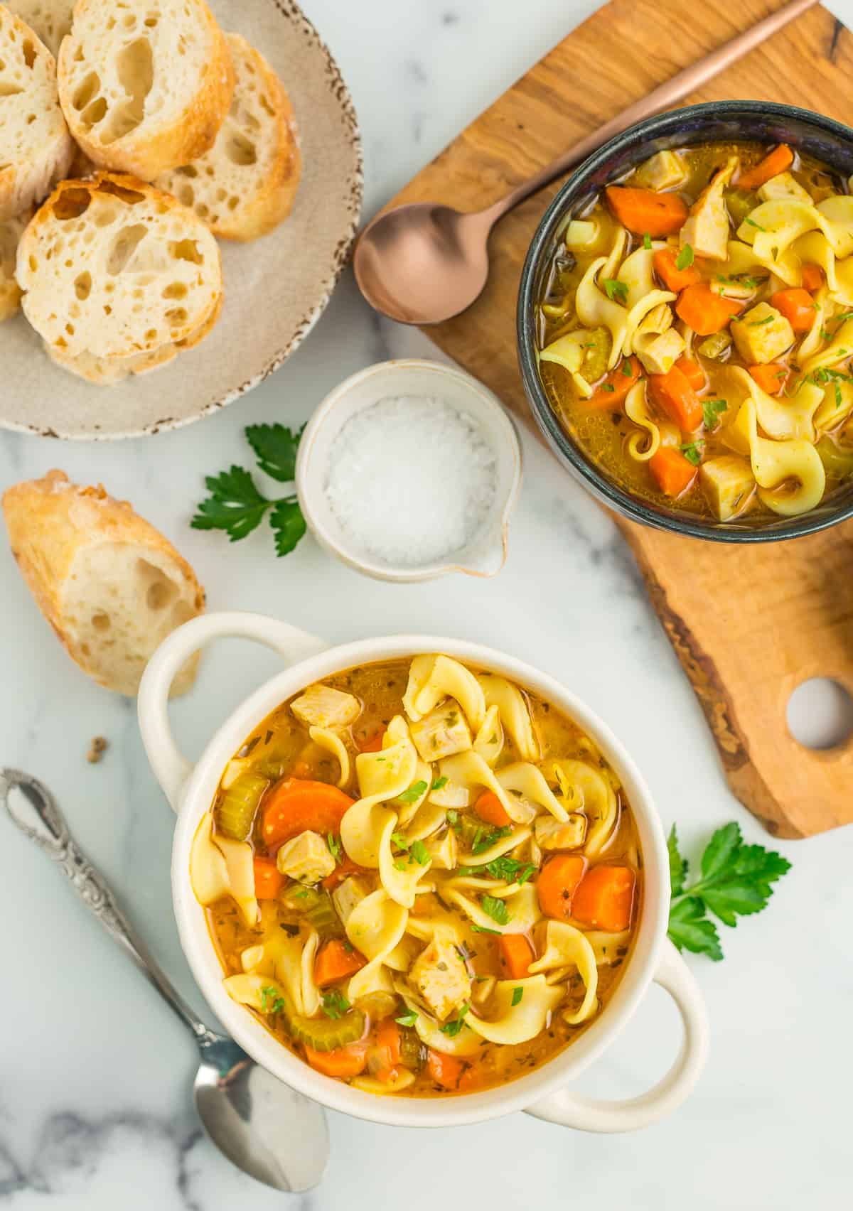 Two bowls of turkey and vegetable noodle soup on a marbled board with baguettes, salt, and other ingredients.