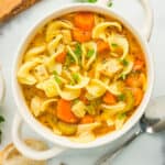 a white bowl of turkey noodle soup topped with chopped parsley. With a spoon, baguette, and cutting board.