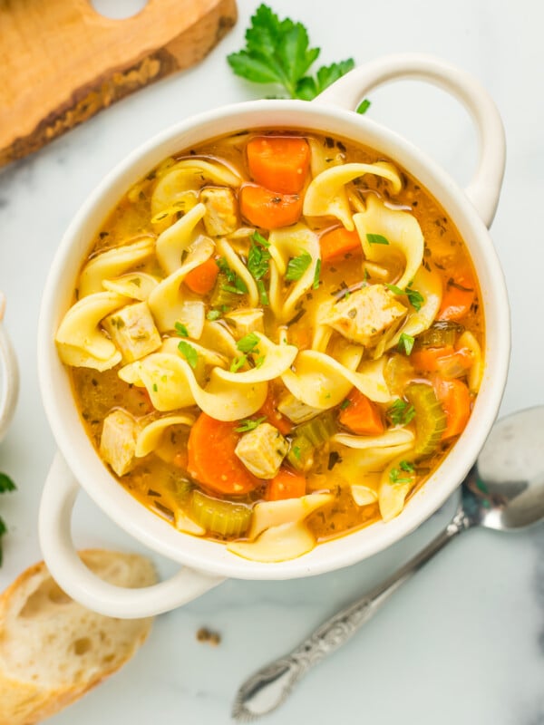 a white bowl of turkey noodle soup topped with chopped parsley. With a spoon, baguette, and cutting board.