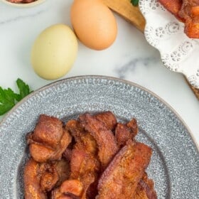 crispy air fryer bacon on a grey plate with a white plate with bacon, eggs, and parsley on a marbled board.