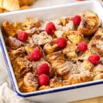 a white casserole dish with croissant bread pudding topped with powdered sugar and raspberries.